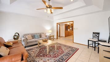 A spacious living room with a high ceiling, a ceiling fan, a large rug, a sofa, a coffee table, and a dining area with chairs.