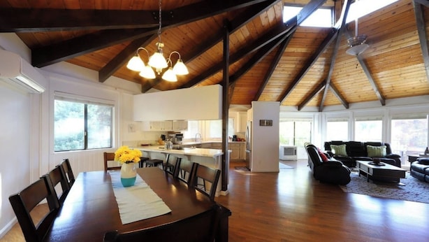 A spacious living area with a wooden ceiling, a dining table with chairs, a kitchen in the background, and a large window providing natural light. - Outdoor Deck • Disc/Pool Table • Fireplace • Grill (Camino)