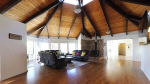 A spacious living room with a high wooden ceiling, a stone fireplace, and a large window. - Outdoor Deck • Disc/Pool Table • Fireplace • Grill (Camino)