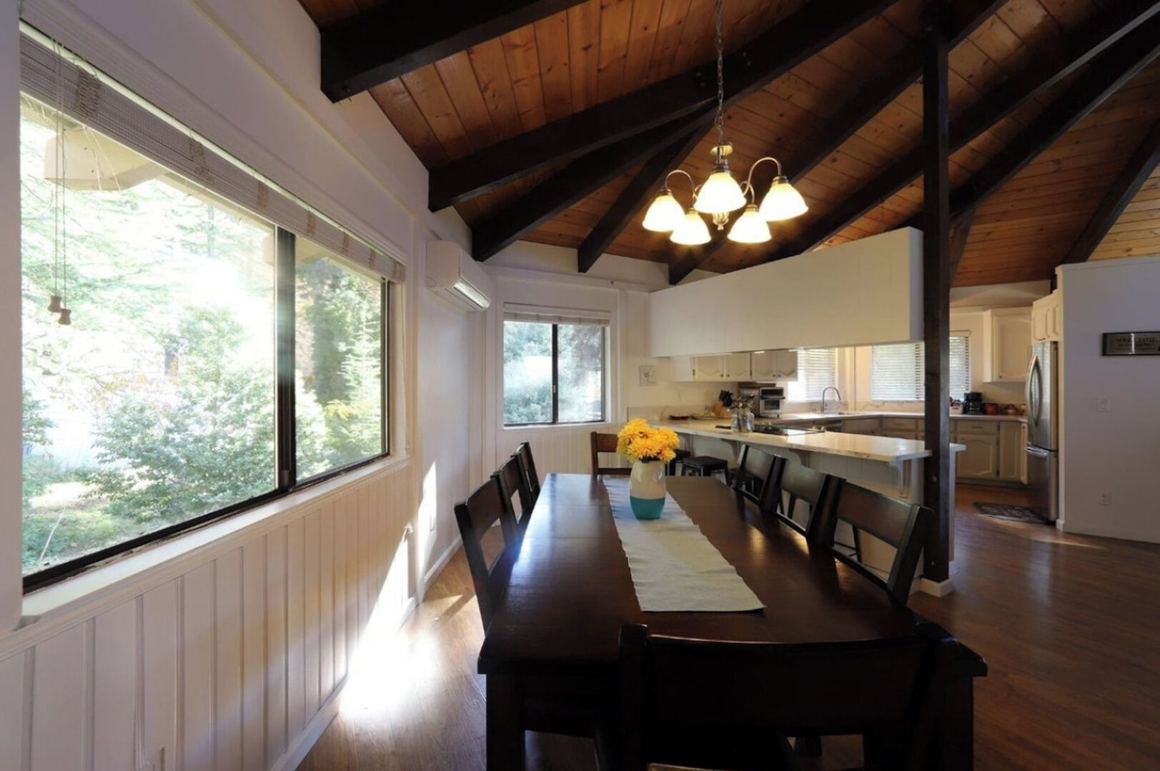 Un comedor con una mesa larga, sillas y un florero. Hay ventanas con persianas, una cocina al fondo y un techo de madera.