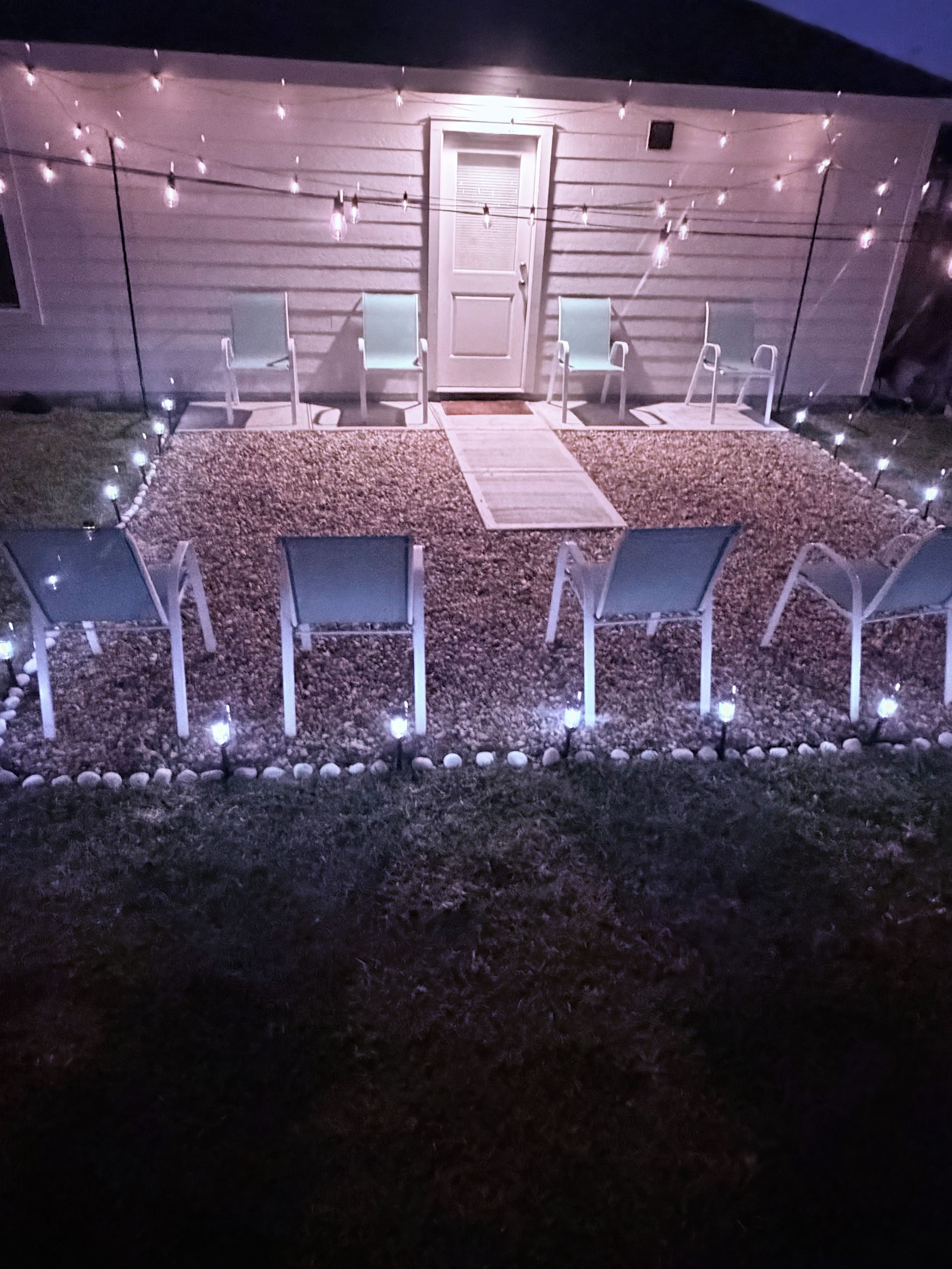 An outdoor seating area with chairs and a table, surrounded by a gravel path and string lights.