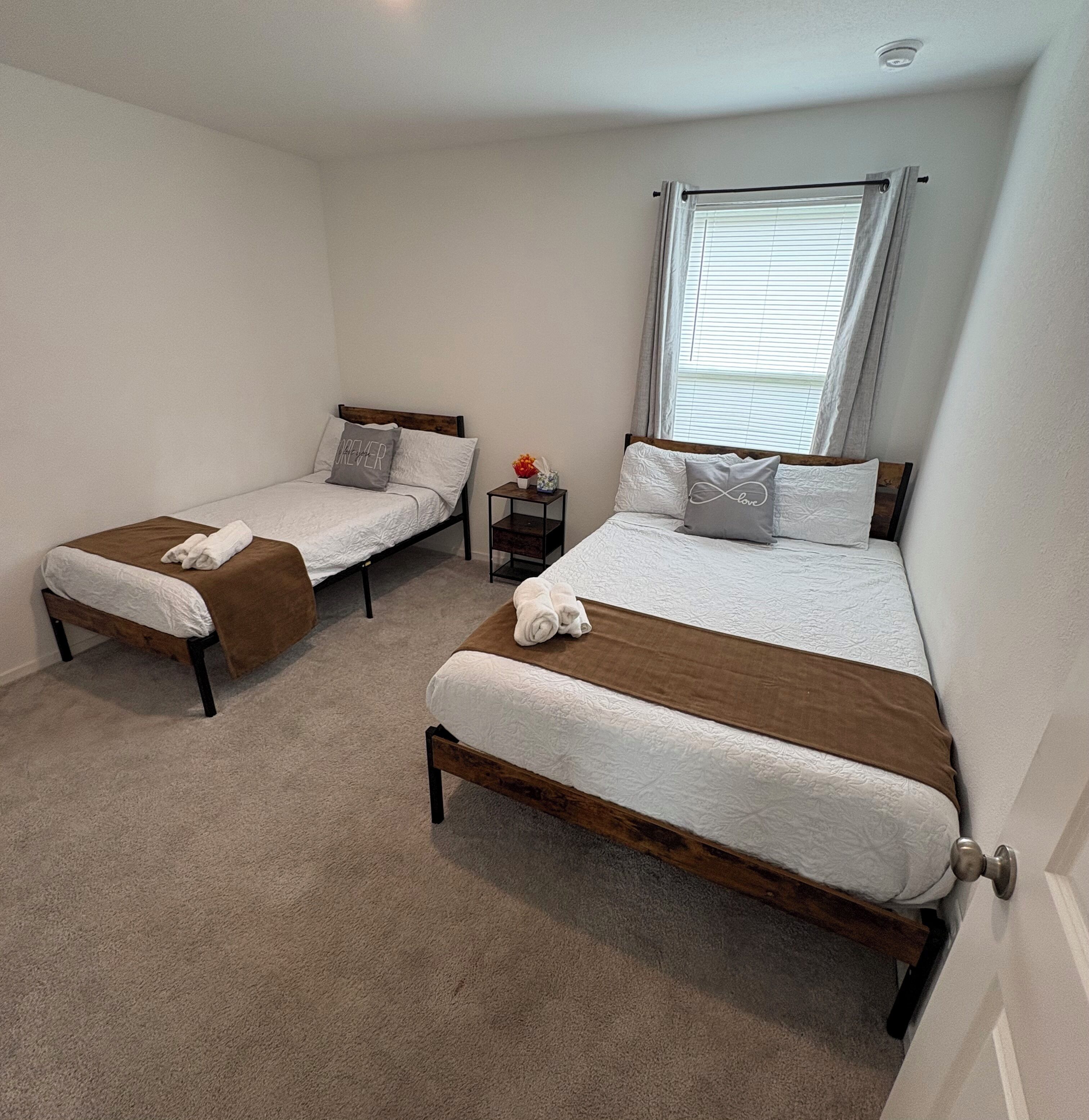 A bedroom with two single beds, a nightstand with a vase, and a window with blinds.