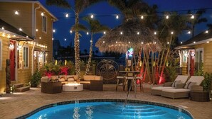 A swimming pool with lounge chairs and a fire pit, surrounded by palm trees and string lights.
