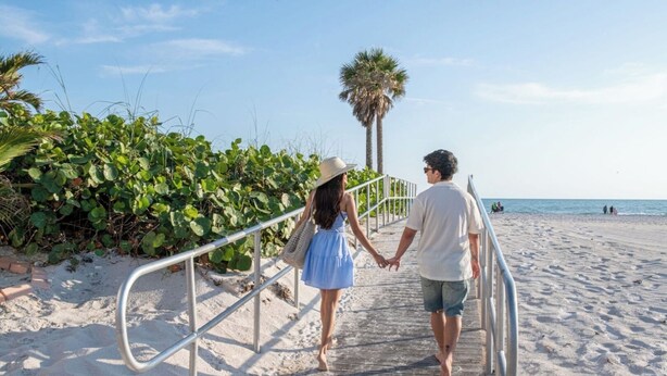 A wooden boardwalk leading to a sandy beach with palm trees and greenery.
