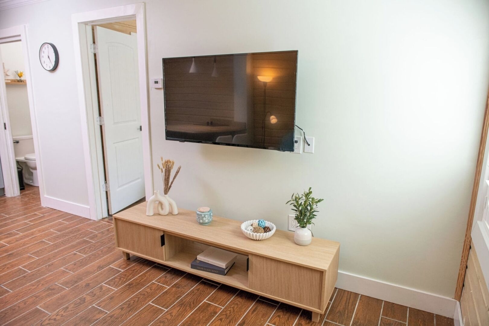 A modern living room with a flat-screen TV mounted on the wall, a wooden console table with decorative items, and a visible door leading to another room.