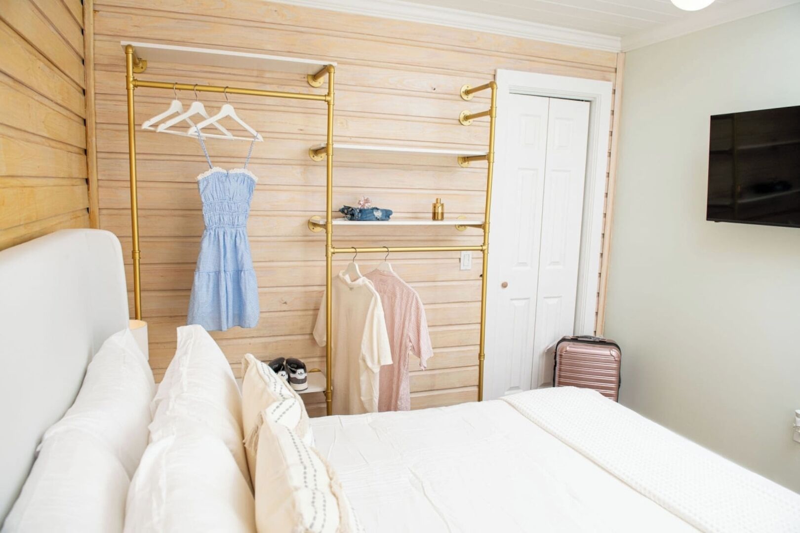 A hotel room with a bed, a wardrobe with hanging clothes, a TV on the wall, and a suitcase.