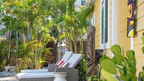 Un área de estar al aire libre iluminada por el sol, con mobiliario de mimbre y una mesa blanca, rodeada de plantas tropicales y un poste de madera decorativo.