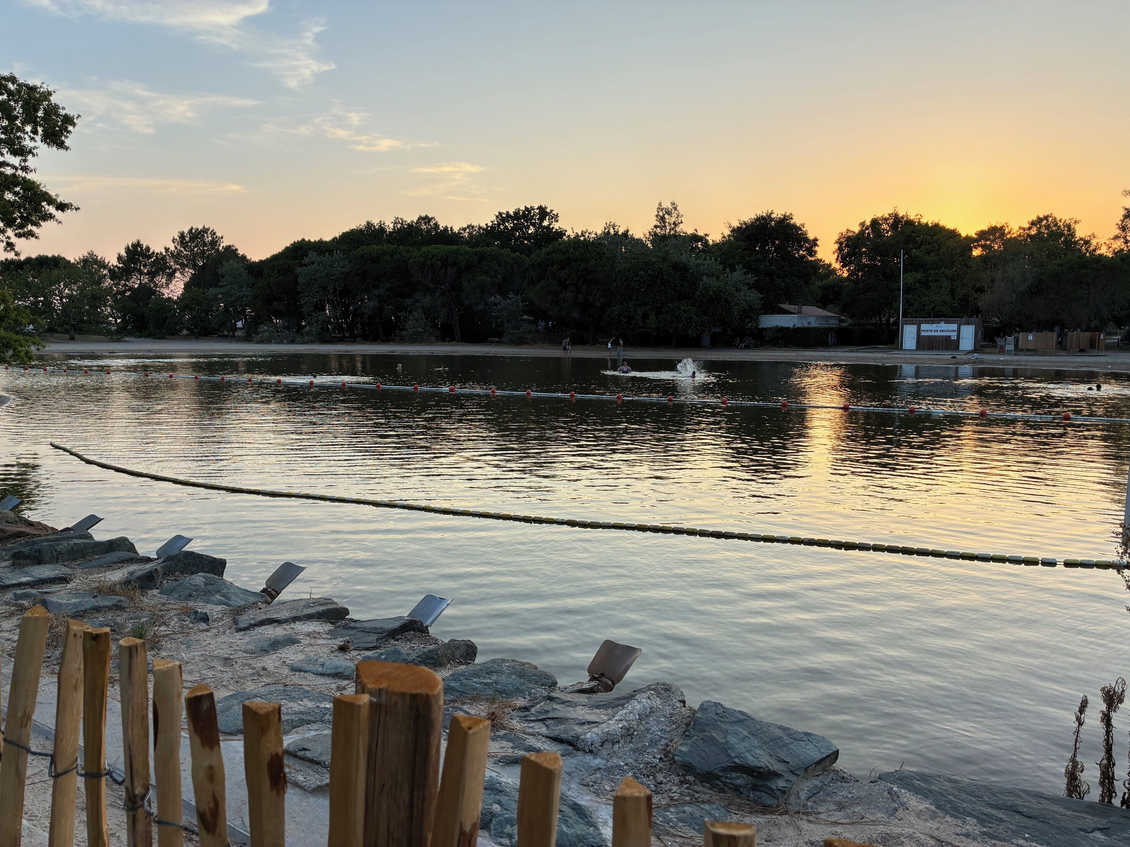 A calm lake with a wooden fence in the foreground and trees in the background.