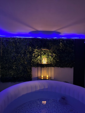 A room with a white circular seating area, a wall with dense foliage, and a table with a lamp.