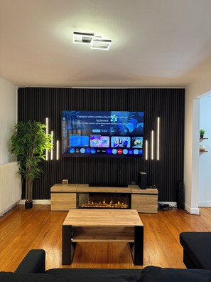 Un saló modern amb televisió de pantalla plana penjada a una paret negra, terres de fusta, una llar de foc central i una planta en test.