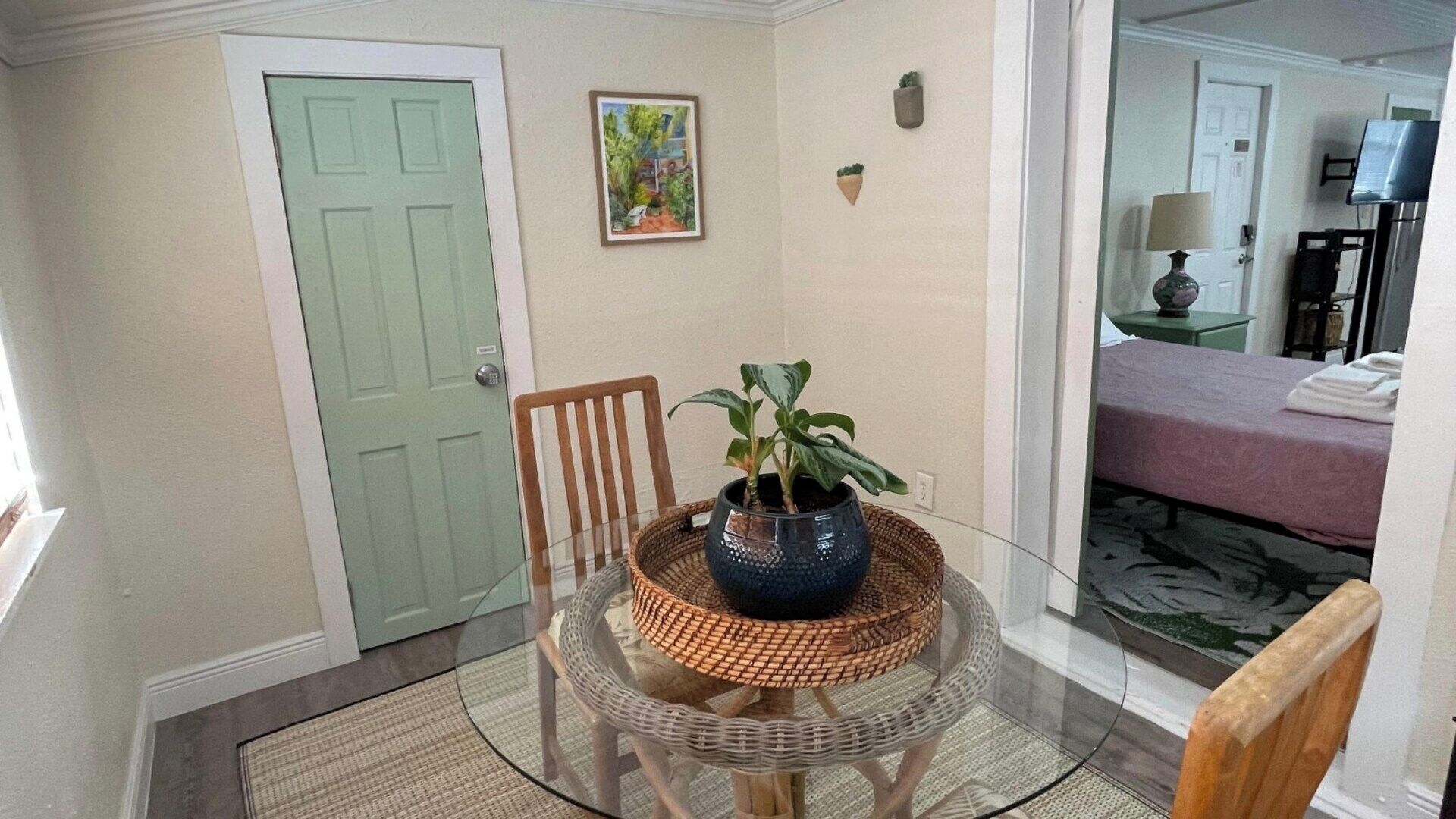 Une pièce comprenant une table à manger, des chaises, une plante en pot, un tableau et un lit recouvert d’une couverture rose.