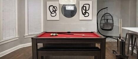 A pool table in a room with a modern design, featuring a hanging light fixture and abstract wall art.