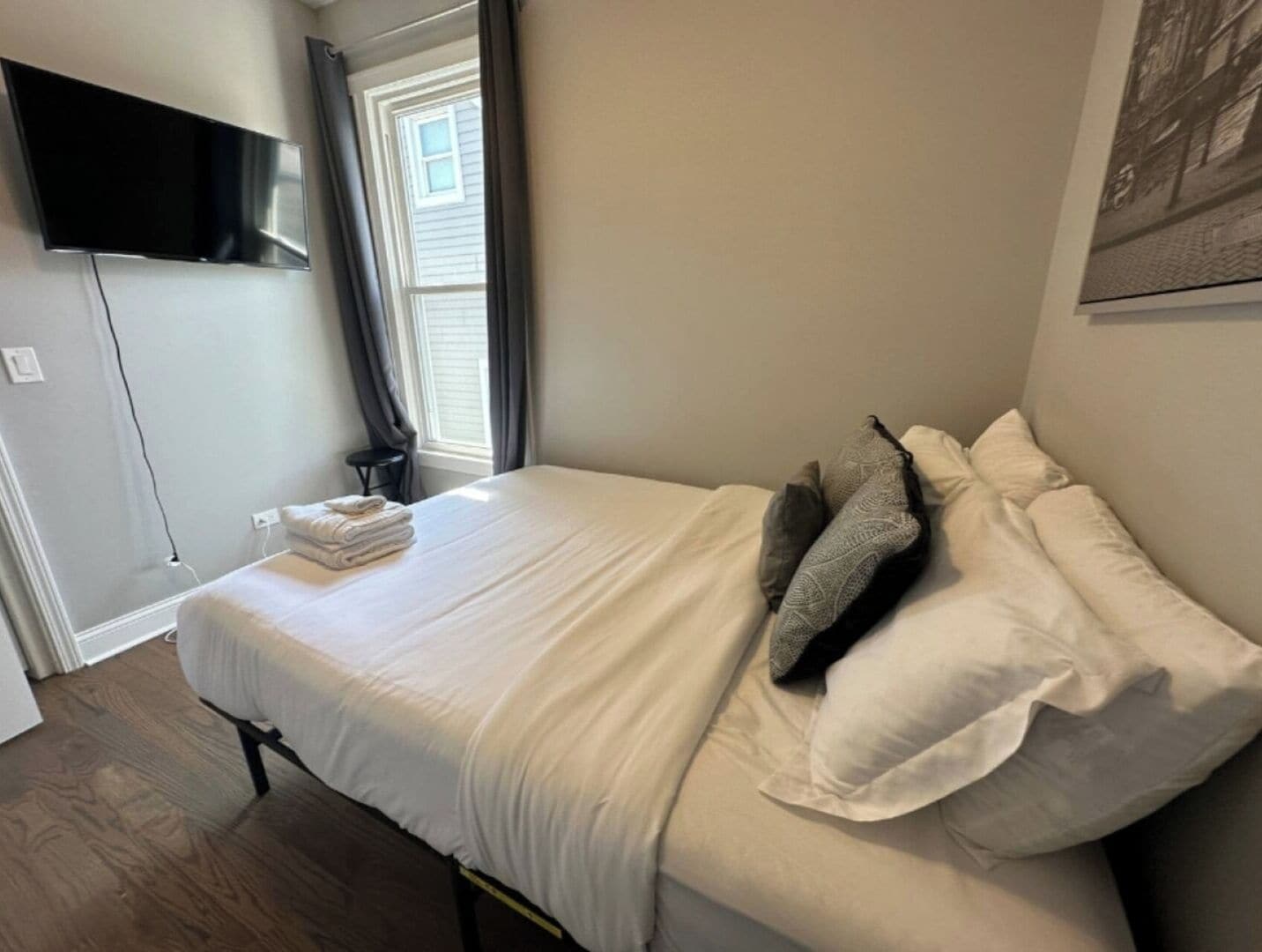 Un lit bien fait, avec du linge de lit blanc et quelques coussins décoratifs, un téléviseur à écran plat fixé au mur et une fenêtre avec des rideaux.