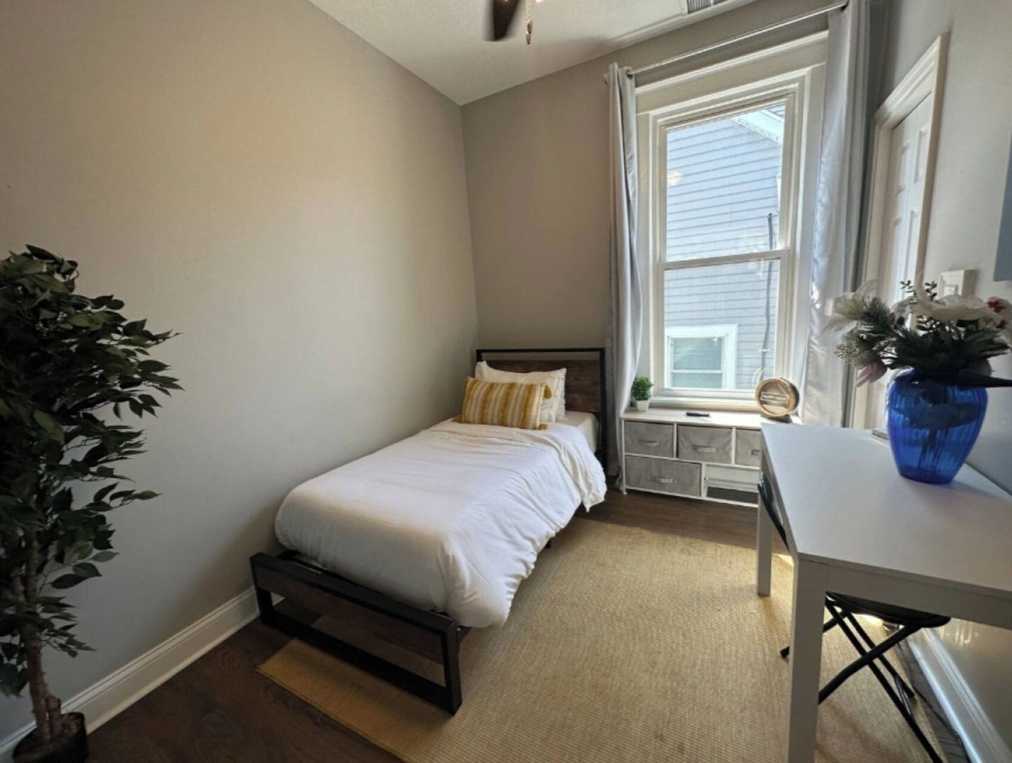 A bedroom with a single bed, a window with curtains, a bedside table with a vase, and a potted plant.