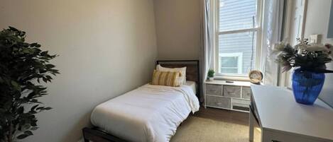 A bedroom with a single bed, a window with curtains, a bedside table with a vase, and a potted plant.