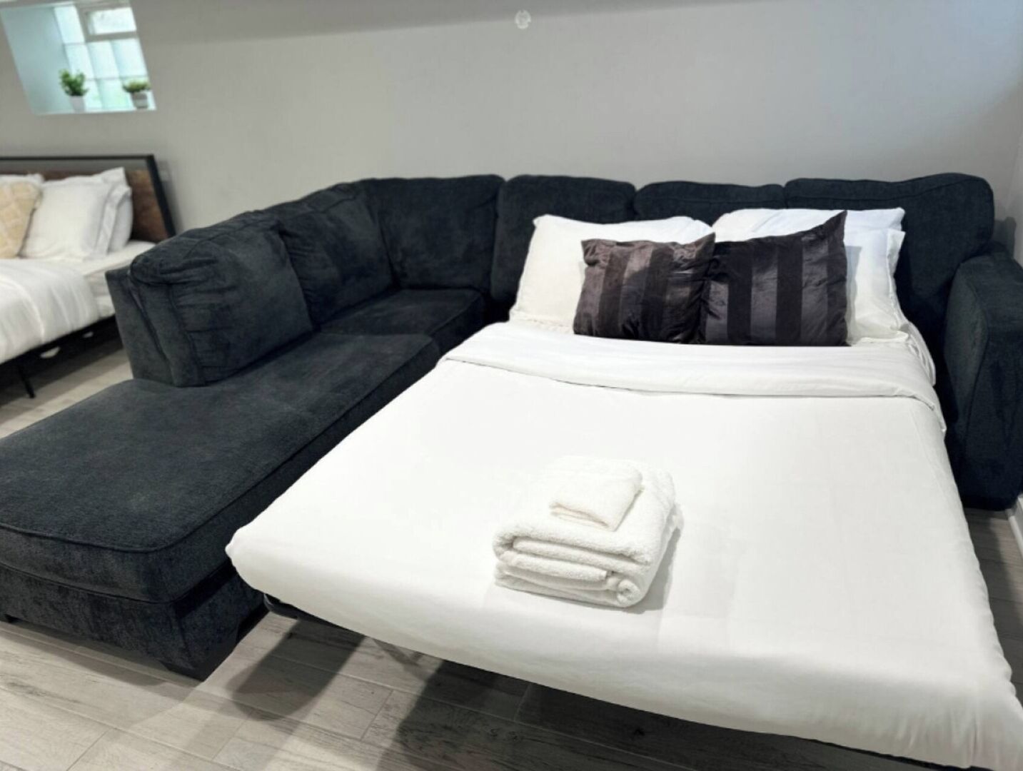 Ein modernes Schlafzimmer mit einer dunkelgrauen Schlafcouch, einem separaten weißen Bett und einem kleinen Nachttisch mit einem zusammengefalteten weißen Handtuch.