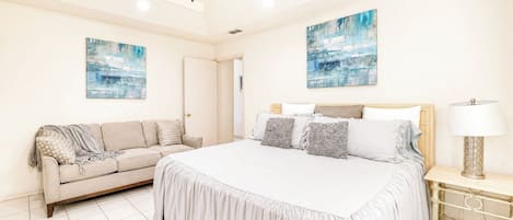 A bedroom with a bed, a sofa, two paintings, a ceiling fan, and a bedside lamp.