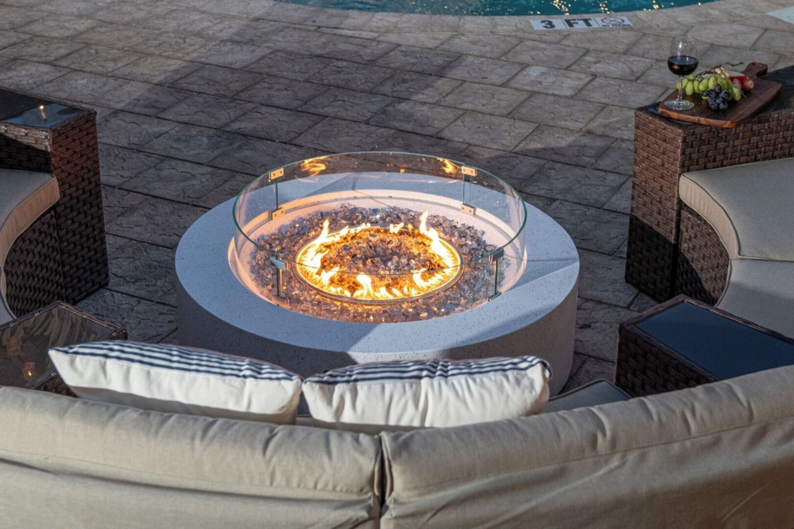A circular outdoor seating area with a glass-top fire pit surrounded by wicker furniture and cushions.