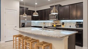 Una cocina moderna con armarios oscuros, electrodomésticos de acero inoxidable, una isla grande con taburetes, iluminación colgante y un salpicadero de azulejos.