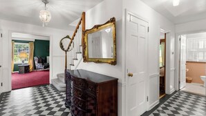 Um corredor espaçoso com chão quadriculado, uma escadaria imponente e um espelho grande com moldura decorada.