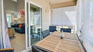 Eine überdachte Terrasse mit einem Holztisch und Stühlen, eine Glastür zu einem Wohnbereich mit einer Couch und Kissen sowie ein Deckenventilator.