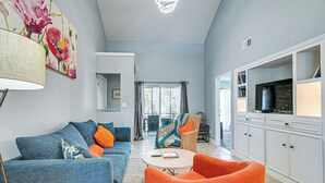Ein Wohnzimmer mit einem blauen Sofa, einem orangefarbenen Sessel und einem Couchtisch. Ein Fernseher steht auf einem Schrank und ein Gemälde hängt an der Wand.