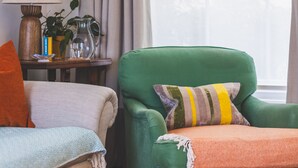 Een kamer met een groene fauteuil, een beige bank, een klein houten bijzettafeltje met een lamp, boeken en een karaf.