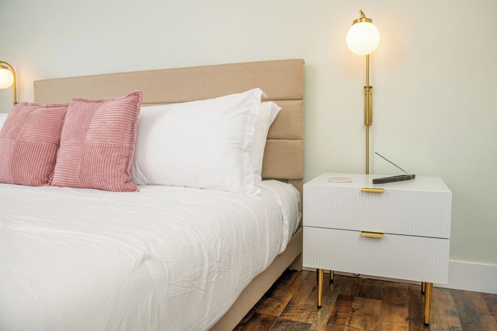 A neatly made bed with white and pink pillows, a beige headboard, and a bedside table with a lamp.