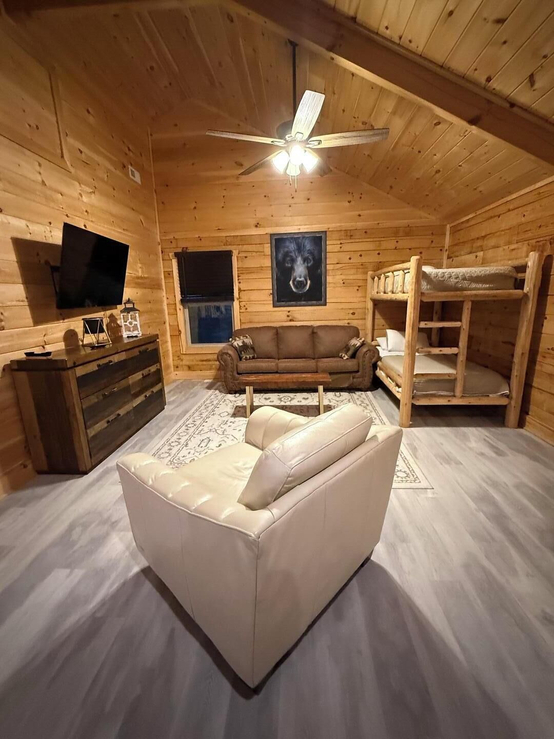 A cozy cabin interior with a wooden ceiling, a ceiling fan, a flat-screen TV, a couch, a wooden bunk bed, and a framed picture on the wall.