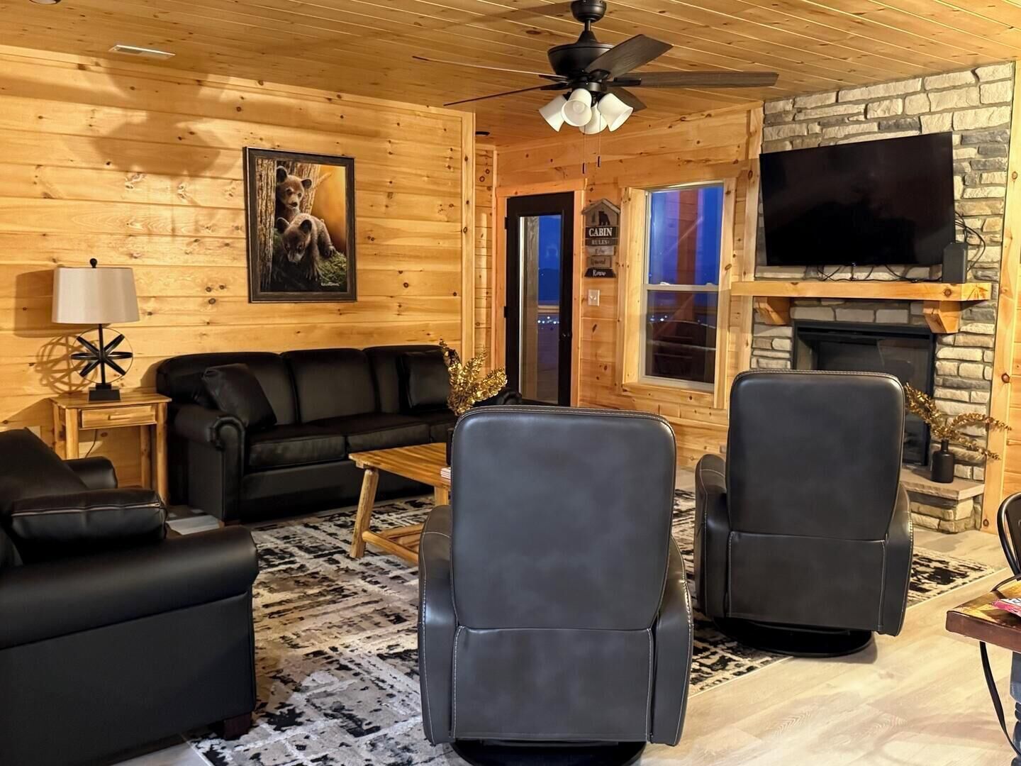 A cozy cabin interior with a fireplace, a black leather sofa, two leather chairs, and a ceiling fan.