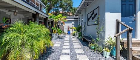 A narrow pathway leading to a building with a blue door, surrounded by potted plants and a small bench.