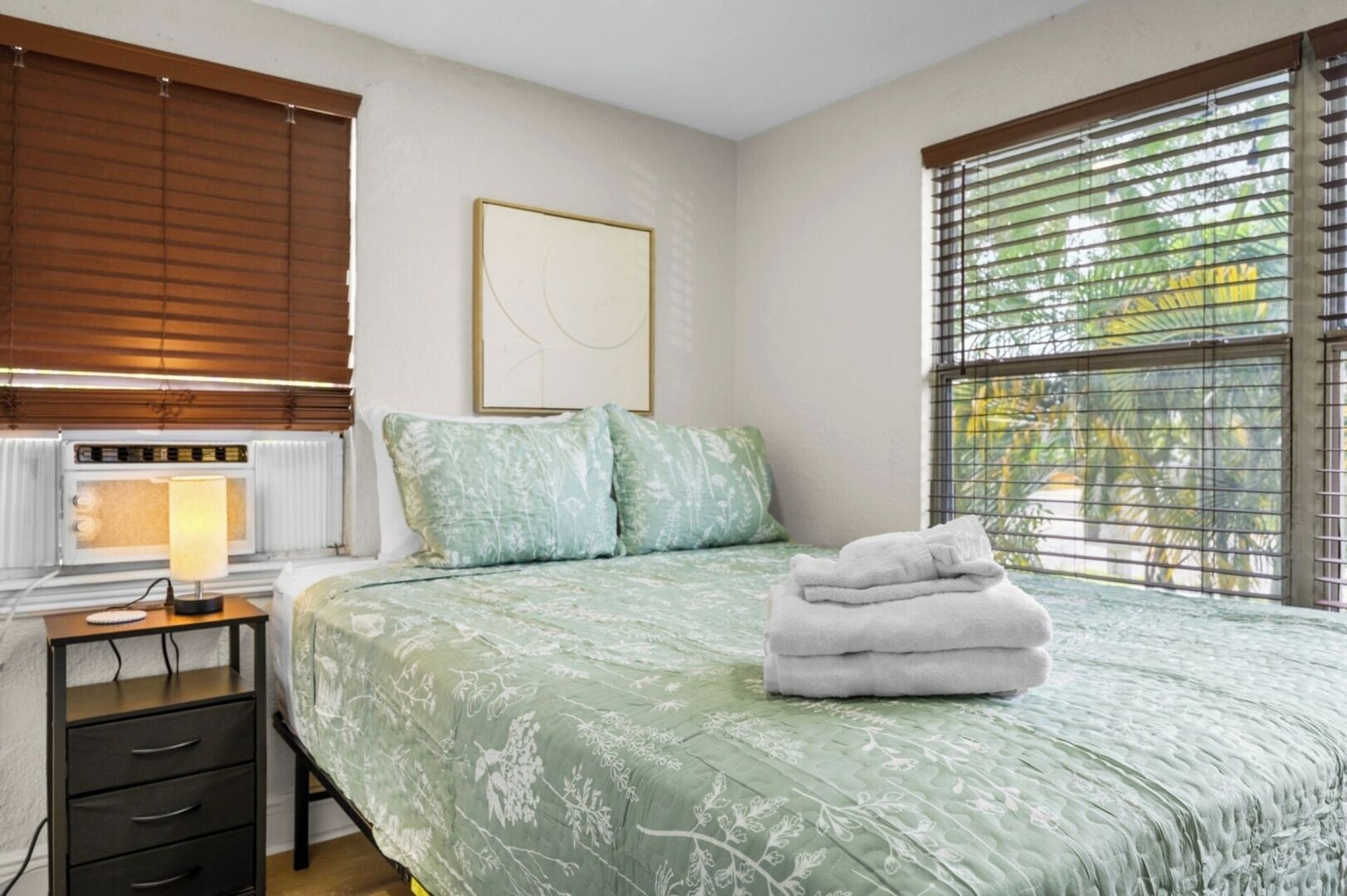 A bedroom with a bed, a bedside table with a lamp, a radiator, and a window with blinds.