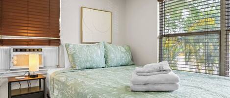 A bedroom with a bed, a bedside table with a lamp, a radiator, and a window with blinds.