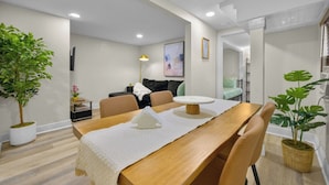A modern dining area with a wooden table, chairs, and a white table runner. There is a potted plant on the left and another on the right. The room has white walls, a ceiling with recessed lighting, and a visible doorway leading to another room. - Spacious Courtyard • Private Deck • Pet Friendly (St. Petersburg)