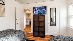 Une pièce avec un téléviseur fixé au-dessus d’un meuble de rangement en osier, une porte donnant sur une autre pièce et une couvre-lit à motifs.
