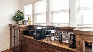 Une cuisine avec un plan de travail en bois, une machine à café, un mixeur et une autre machine à café. On y trouve des fenêtres équipées de stores, une plante en pot et un meuble avec des tiroirs.