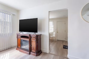 A room with a flat-screen TV on a wooden cabinet, a white door, and a view into another room with a chair and a small table. - Stylish 2BR Patio, BBQ and Parking (Los Angeles)