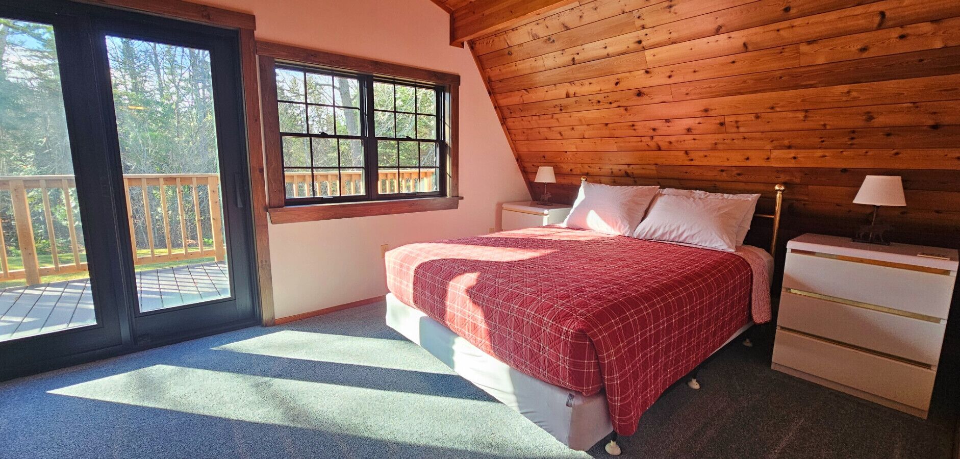 A bedroom with a wooden ceiling, a large window, a bed with a red blanket, a nightstand, and a dresser.