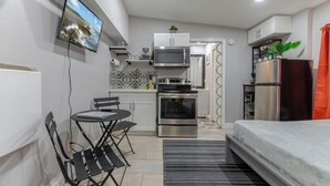 Eine kompakte Küche mit Mikrowelle, Backofen und Kühlschrank, ein kleiner Essbereich mit Tisch und Stühlen sowie ein Bett mit grauer Decke.