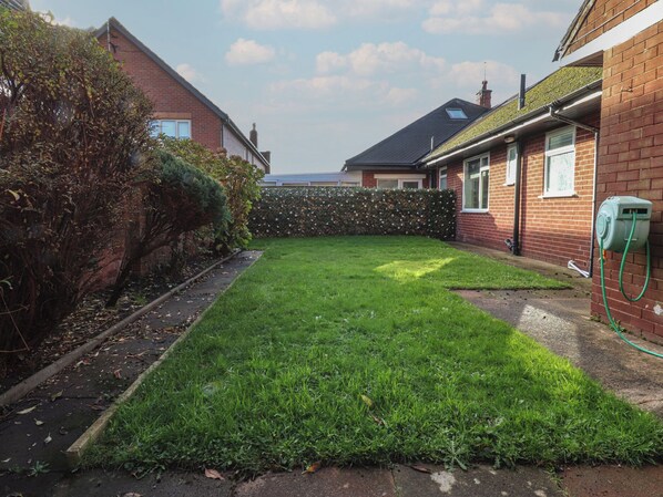 A residential backyard with a well-maintained lawn, a brick house, and a green hose connected to a wall-mounted electrical box.