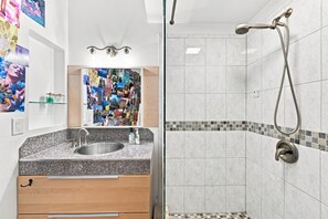 Un baño con lavamanos de encimera de granito, un espejo con un collage de fotografías y una ducha con cabezal de ducha manual.