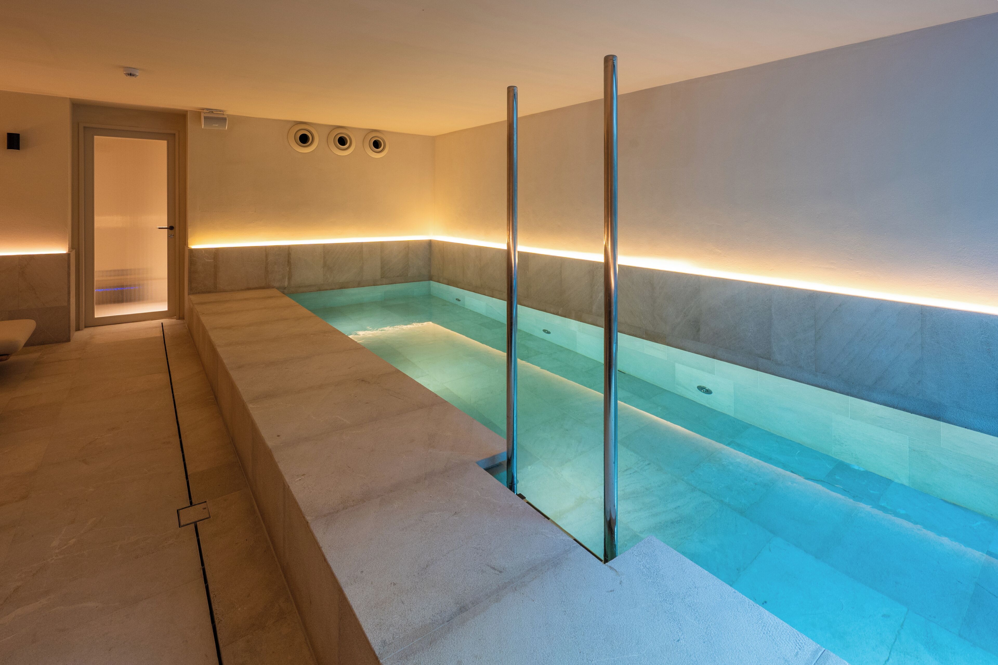 A modern indoor swimming pool with clear glass walls, a tiled floor, and a minimalist design.