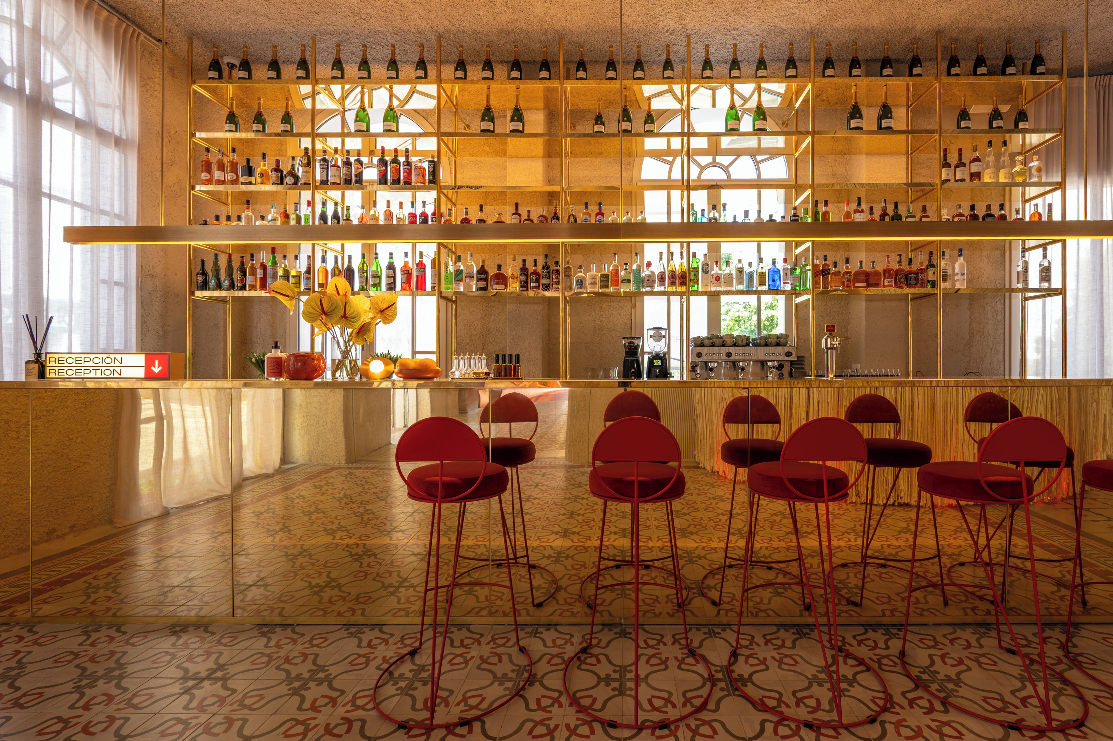 Una zona de bar con estanterías llenas de botellas, un mostrador de recepción y varias sillas rojas.