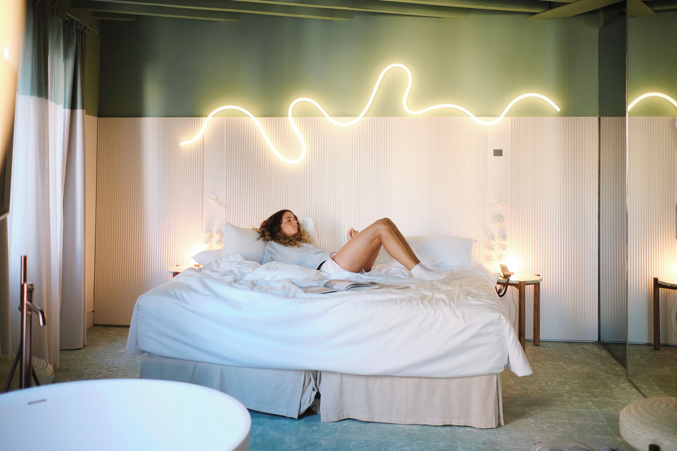 A bed with white bedding, a bedside table with a lamp, and a wall with a light fixture.