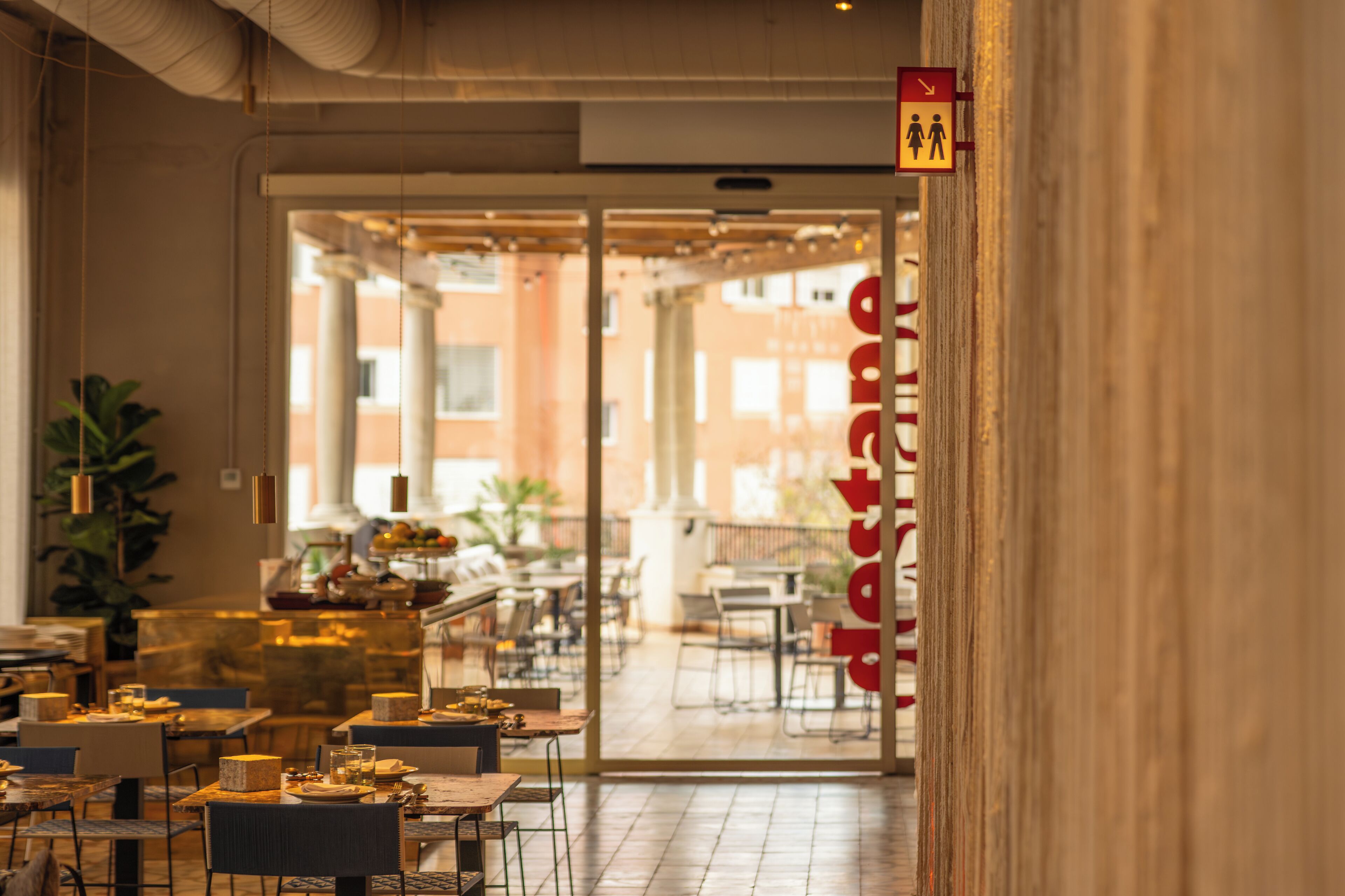 Vista interior de un restaurante con mesas y sillas, una entrada visible y un letrero que indica los baños.