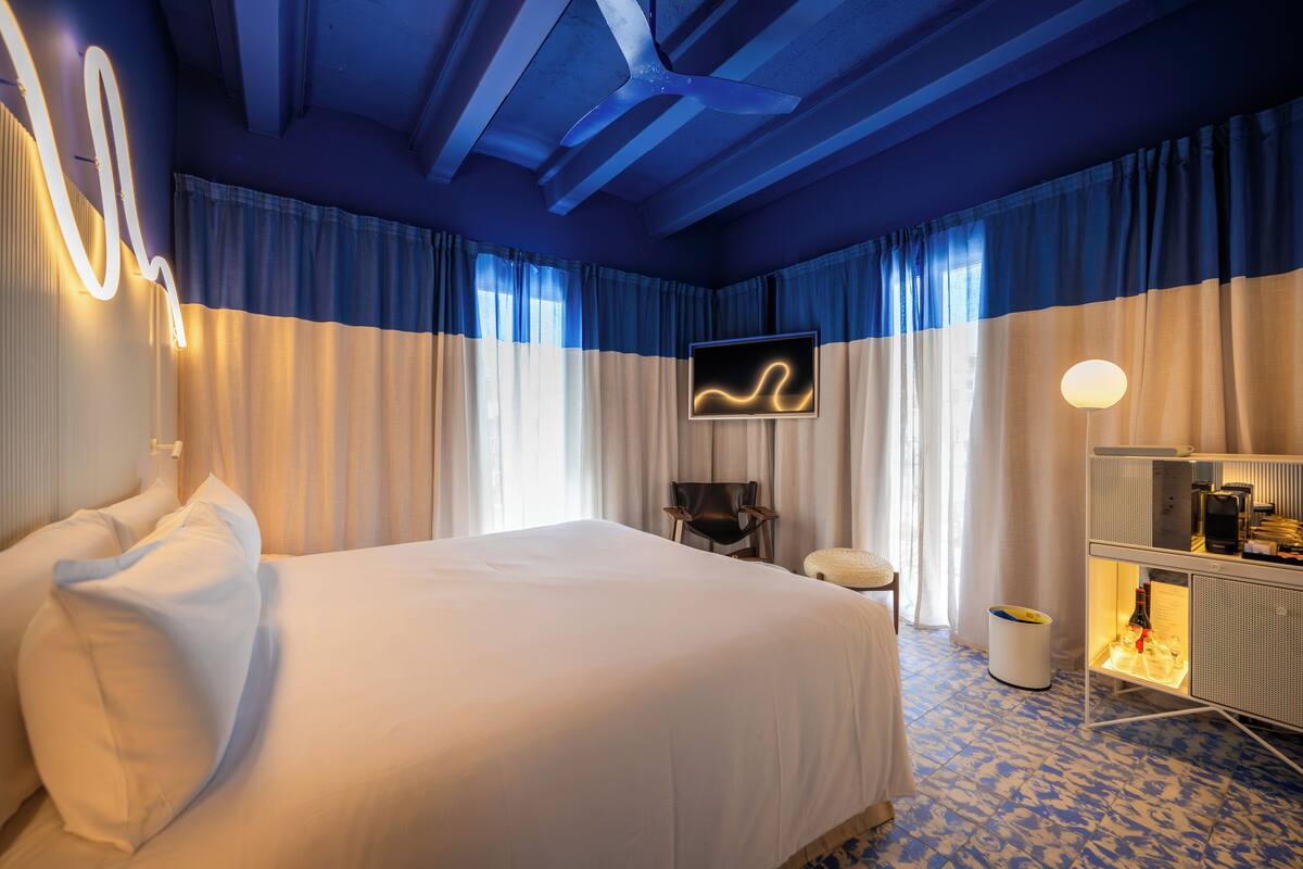 Habitación de hotel con una cama grande, un televisor en la pared, una mesita con una lámpara y un minibar.