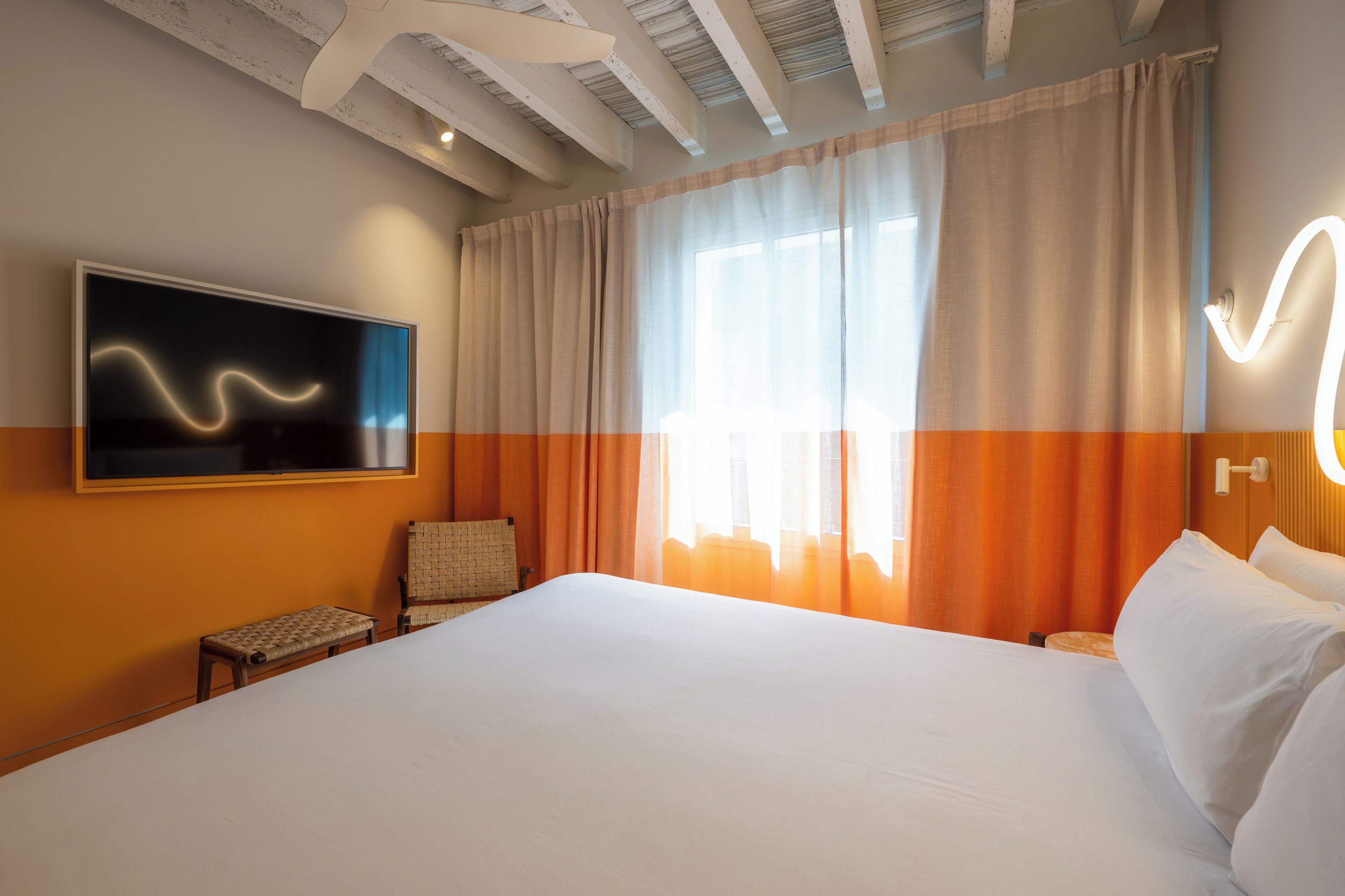 Une chambre d’hôtel avec un grand lit, un téléviseur fixé au mur, une chaise et une fenêtre avec des rideaux.