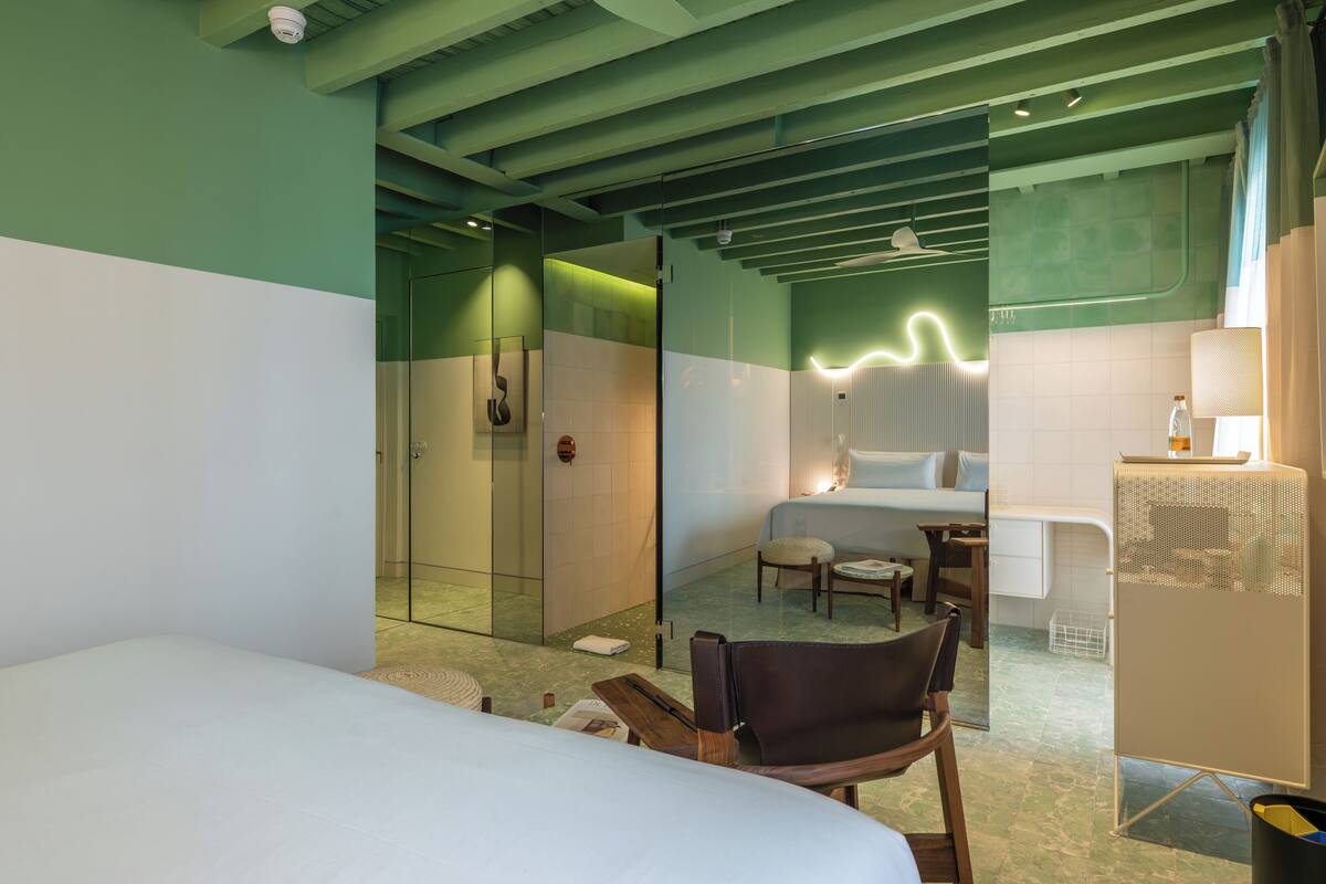 Habitación de hotel con cama, una silla, escritorio, ducha y baño con espejo.