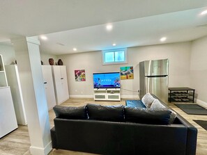A modern living room with a black leather sofa, a flat-screen TV, a stainless steel refrigerator, and a small table with shoes. - Luxurious 2 bedroom Heated floor 2 bathroom near to Chicopee Hill & Airport (Kitchener)