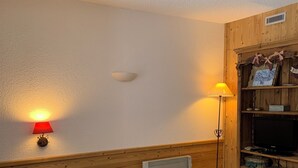 Une pièce confortable avec un lit, une petite table, un radiateur, une lampe et une étagère où sont posés divers objets.